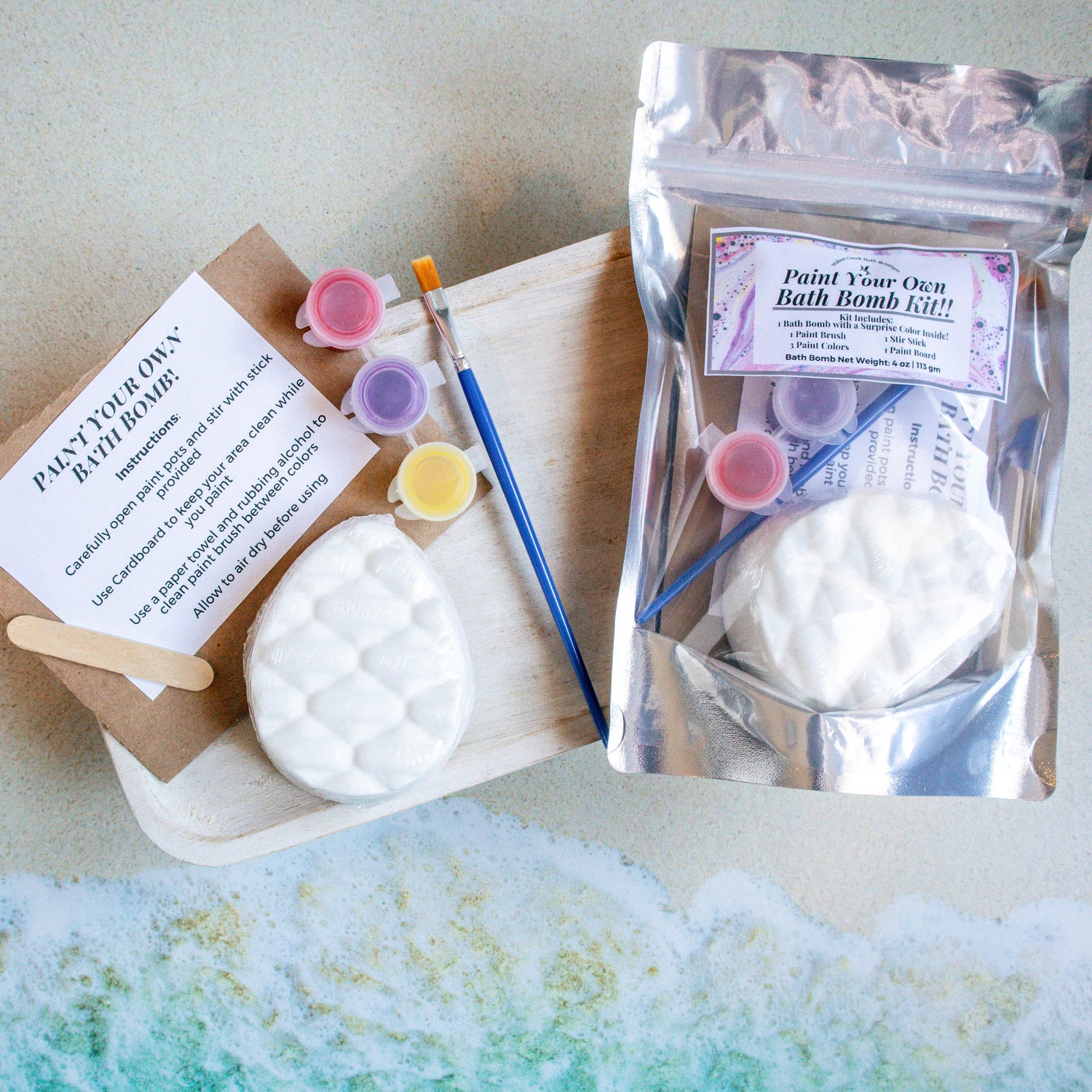 there is a bag of the dino bath bomb kit to the right of the contents splayed out.  the dino egg along with a paint brush, paint pots of red, purple and yellow skin safe colorants as the pain. they are sitting on a piece of cardboard along with the instruction sheet and a stir stick.there is an image of waves crashing on the bottom of the frame.