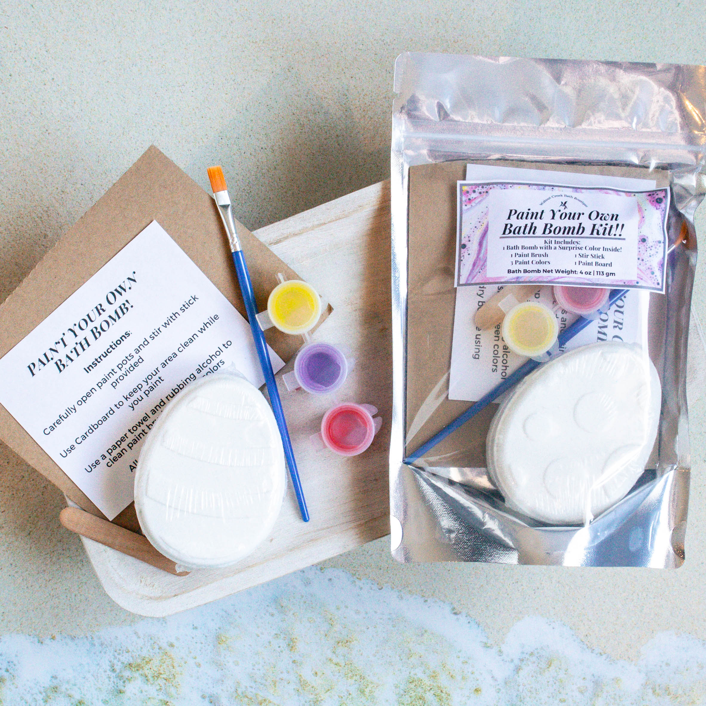 there is a bag of the easter bath bomb kit to the right of the contents splayed out. the Easter egg along with a paint brush, paint pots of red, purple and yellow skin safe colorants as the pain. they are sitting on a piece of cardboard along with the instruction sheet and a stir stick.there is an image of waves crashing on the bottom of the frame.