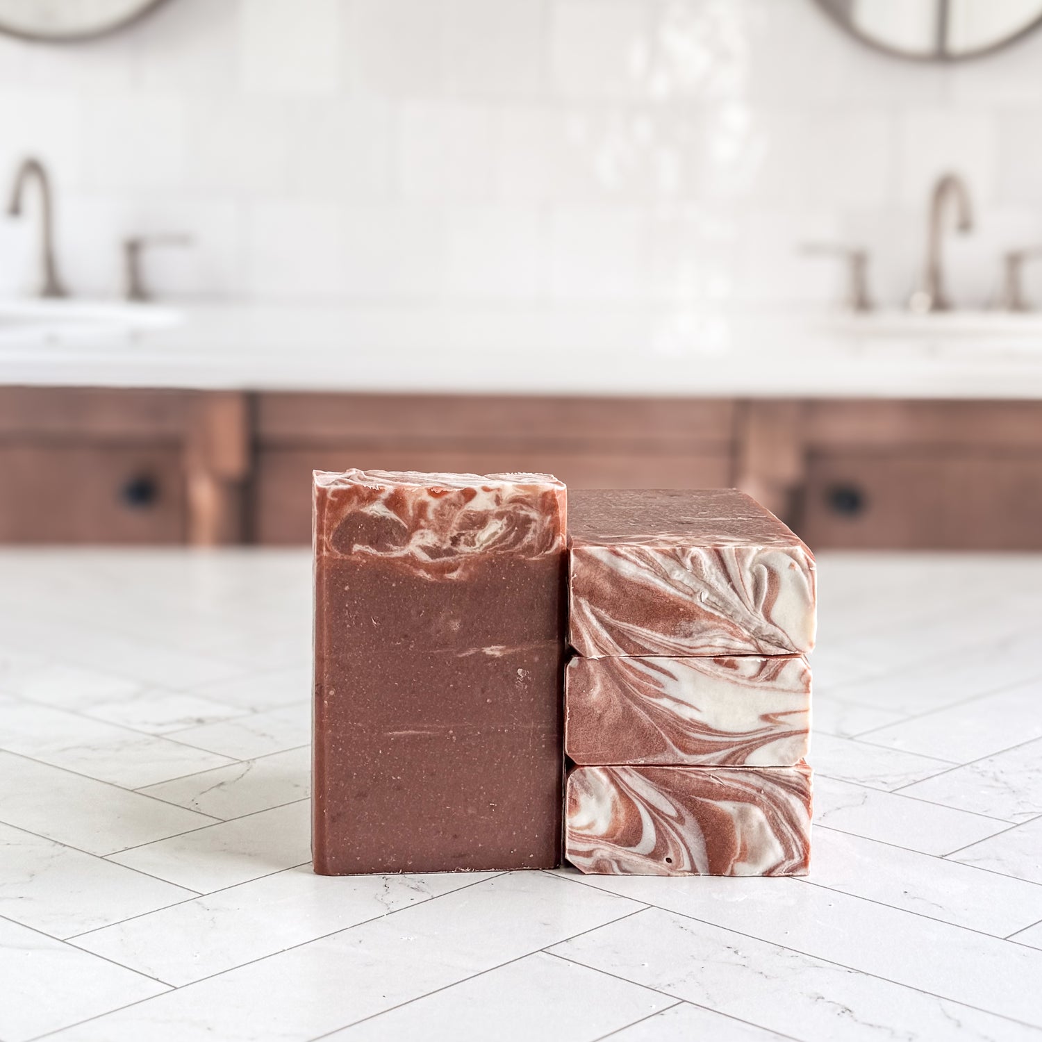 Two handmade soap bars standing upright on a white tiled countertop in a bathroom setting. One bar is solid medium brown, and the other is stacked with a marbled swirl pattern of brown, cream, and pinkish tones.