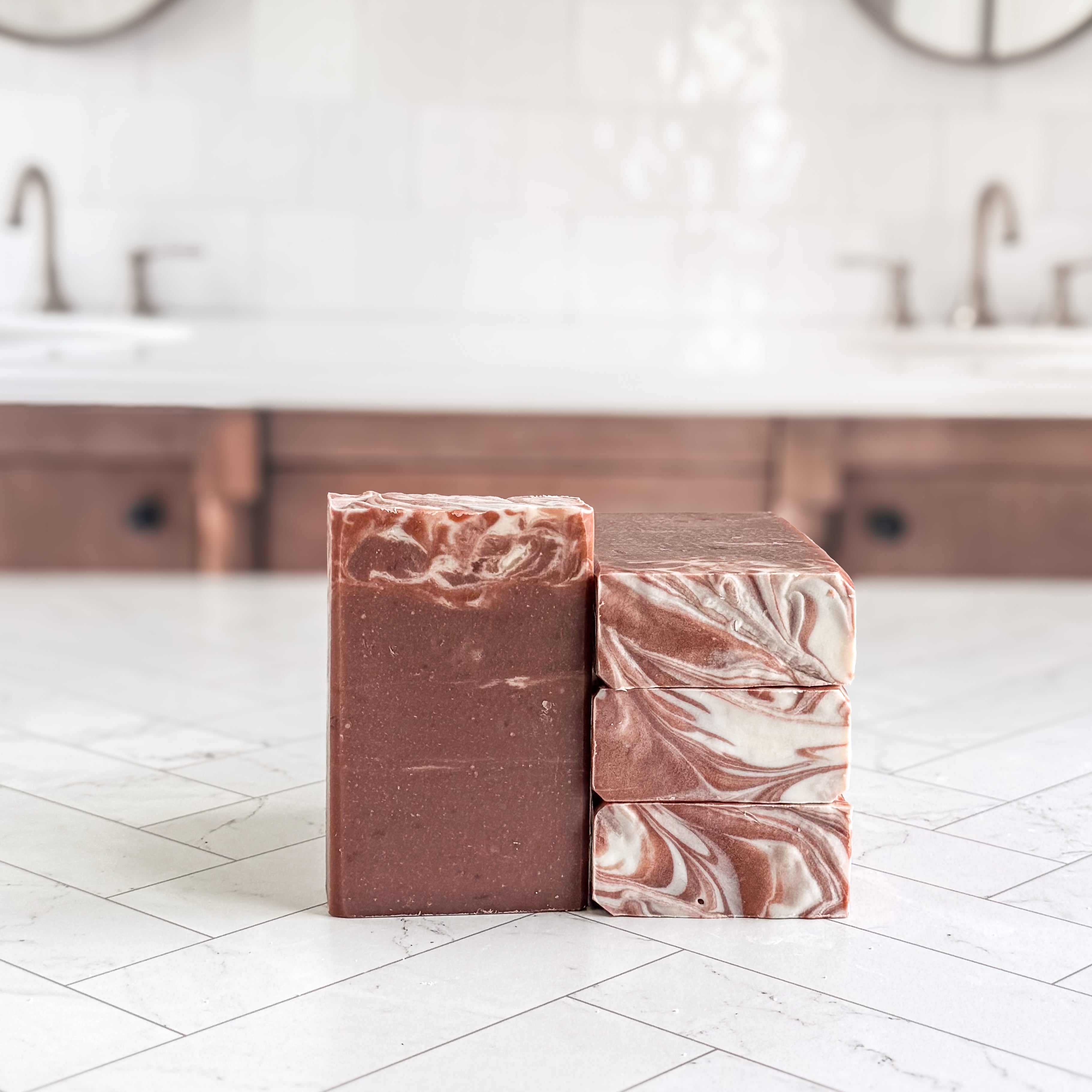 Two handmade soap bars standing upright on a white tiled countertop in a bathroom setting. One bar is solid medium brown, and the other is stacked with a marbled swirl pattern of brown, cream, and pinkish tones.