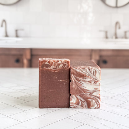 Two handmade soap bars standing upright on a white tiled countertop in a bathroom setting. One bar is solid medium brown, and the other is stacked with a marbled swirl pattern of brown, cream, and pinkish tones.