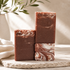 Three rectangular bars of handmade soap in warm brown tones with marbled swirls, arranged on a stone slab with a white vase and soft sunlight in the background.