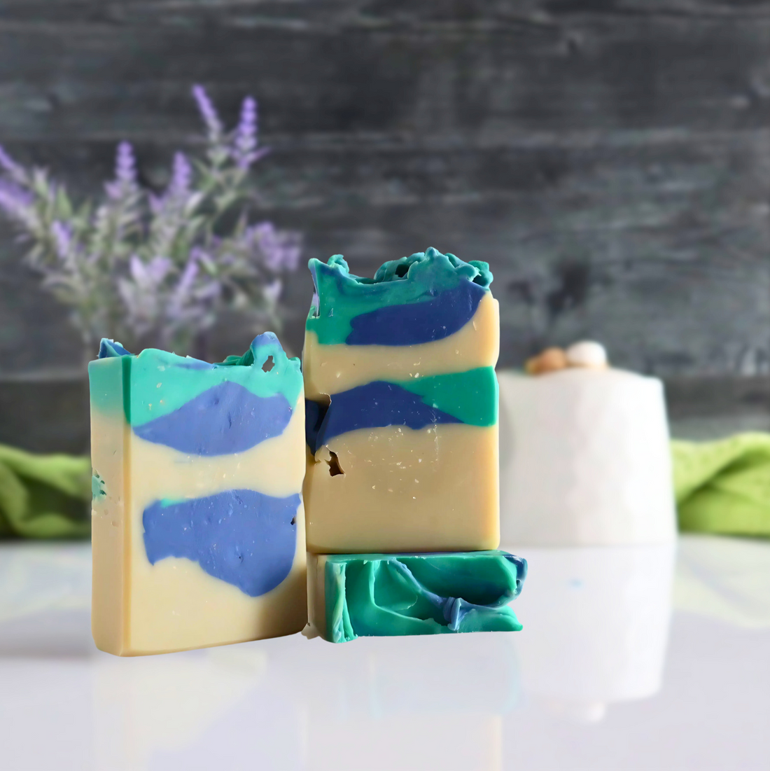 3 patchouli rain soaps are shown, they are a creamy white with thick blue and green patches layered in. they are sitting on a white base with a black background