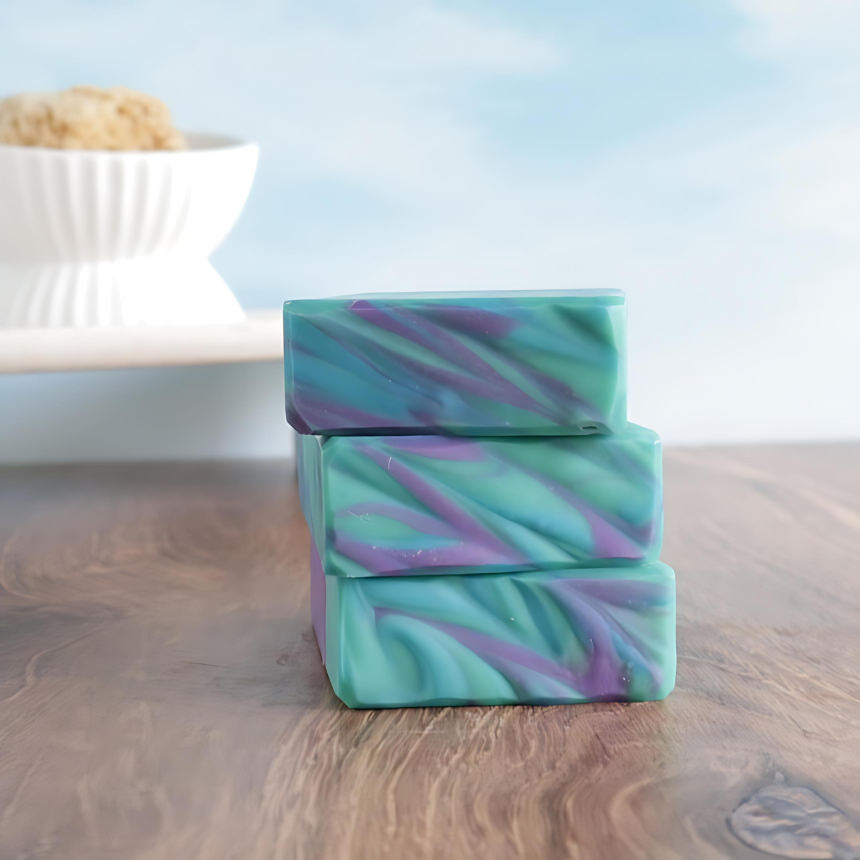 3 patchouli rain soaps are shown stacked on top of each other showing the textured tops with the blue, purple and green swirls.  in the background is a blue sky with whispy clouds. behind the soaps is another tray with a white soap dish with some loofah peeking out of it.