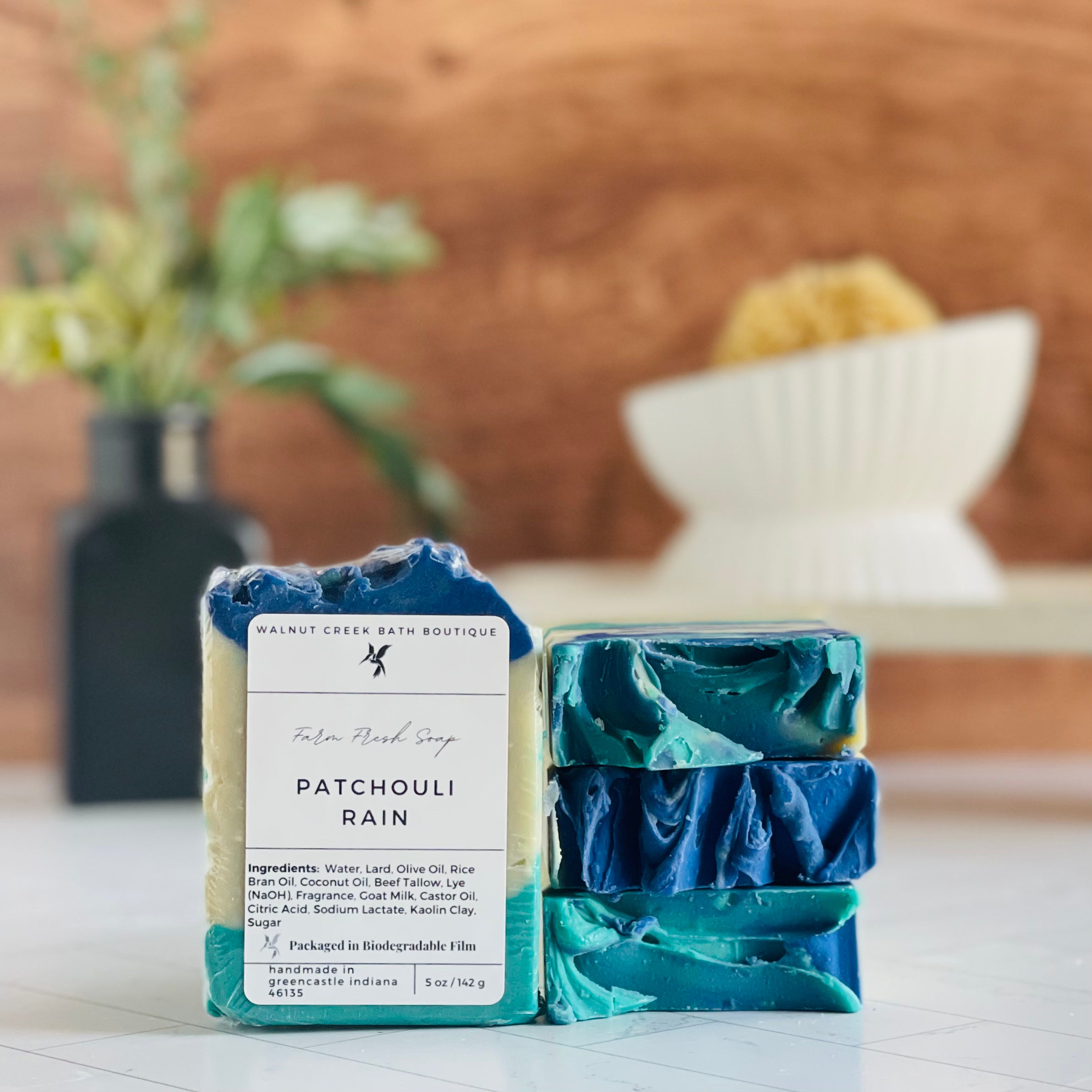 patchouli rain soaps are shown, one standing facing out to show the label. Next to this are three more soaps stacked on top to show the sculpted tops. in the background is a soap dish on a stand and a small vase with some greenery.