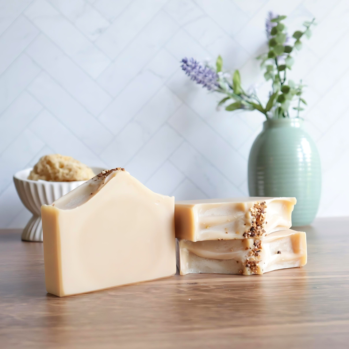 3 Patchouli Sunrise soaps are shown, 1 is standing to the left of the other 2. one of those is laying flat to show sculpted top with orange granules on top  and there is another one standing on top of that. in the background is a white dish with a loofah in the left and back right has a sage green vase with lavender buds.