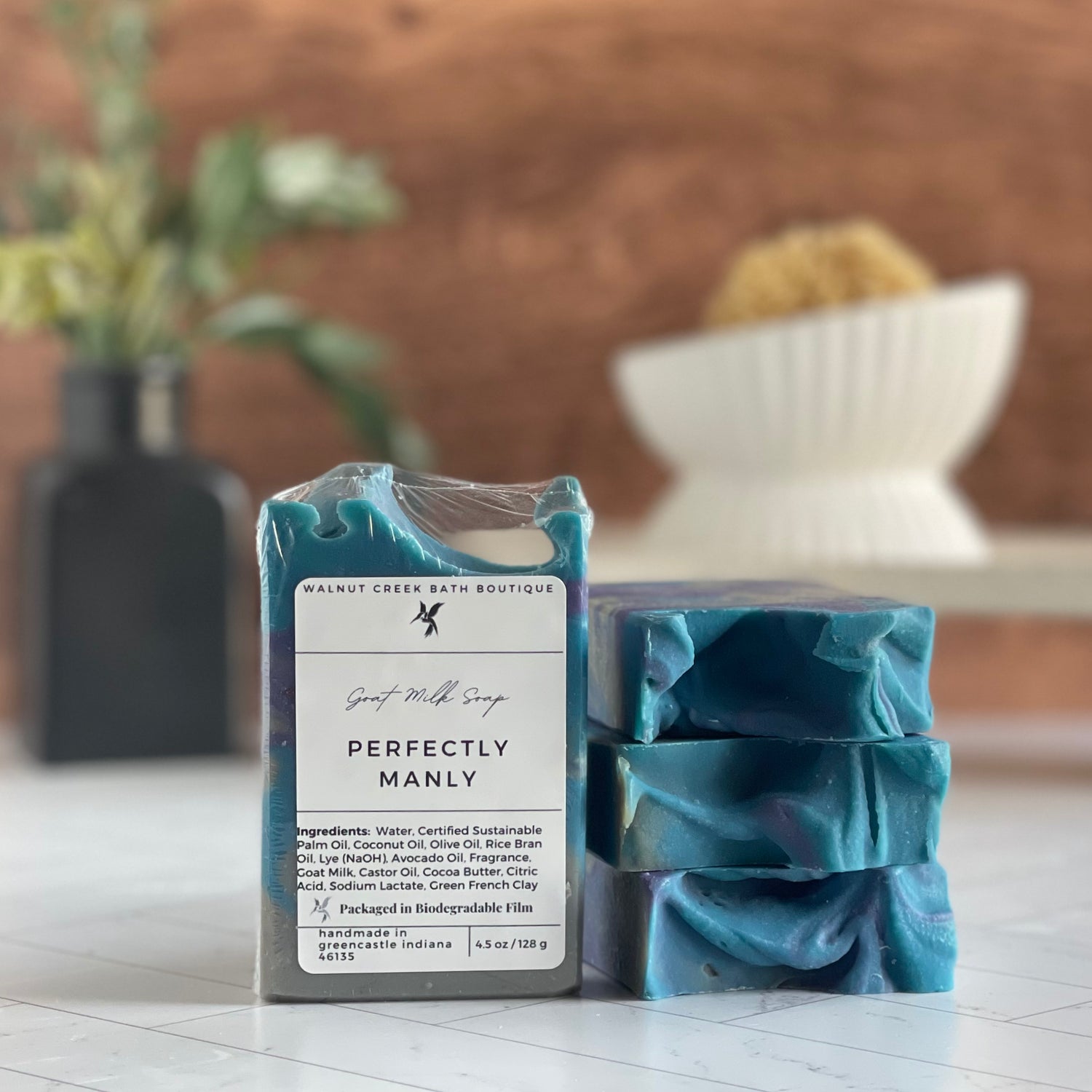 perfectly manly soaps are shown, one standing facing out to show the label. Next to this are three more soaps stacked on top to show the sculpted tops. in the background is a soap dish on a stand and a small vase with some greenery.