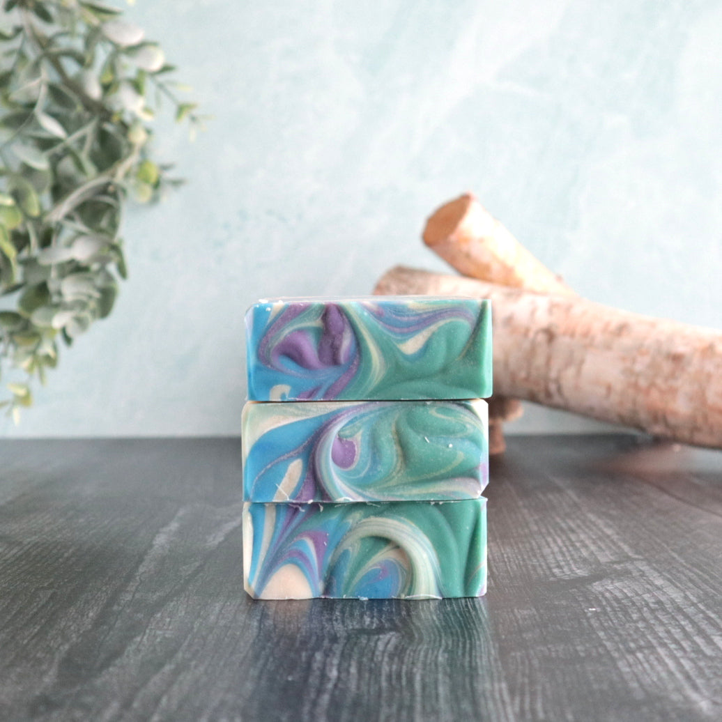 3 bars of perfectly manly soaps are pictured stacked on top of each other to show the pretty swirls on top.  The top has  a white base with blue, green and purple swirls. There is some greenery peaking out in the left and some small logs in the back right. The background is a pretty blue-green color.