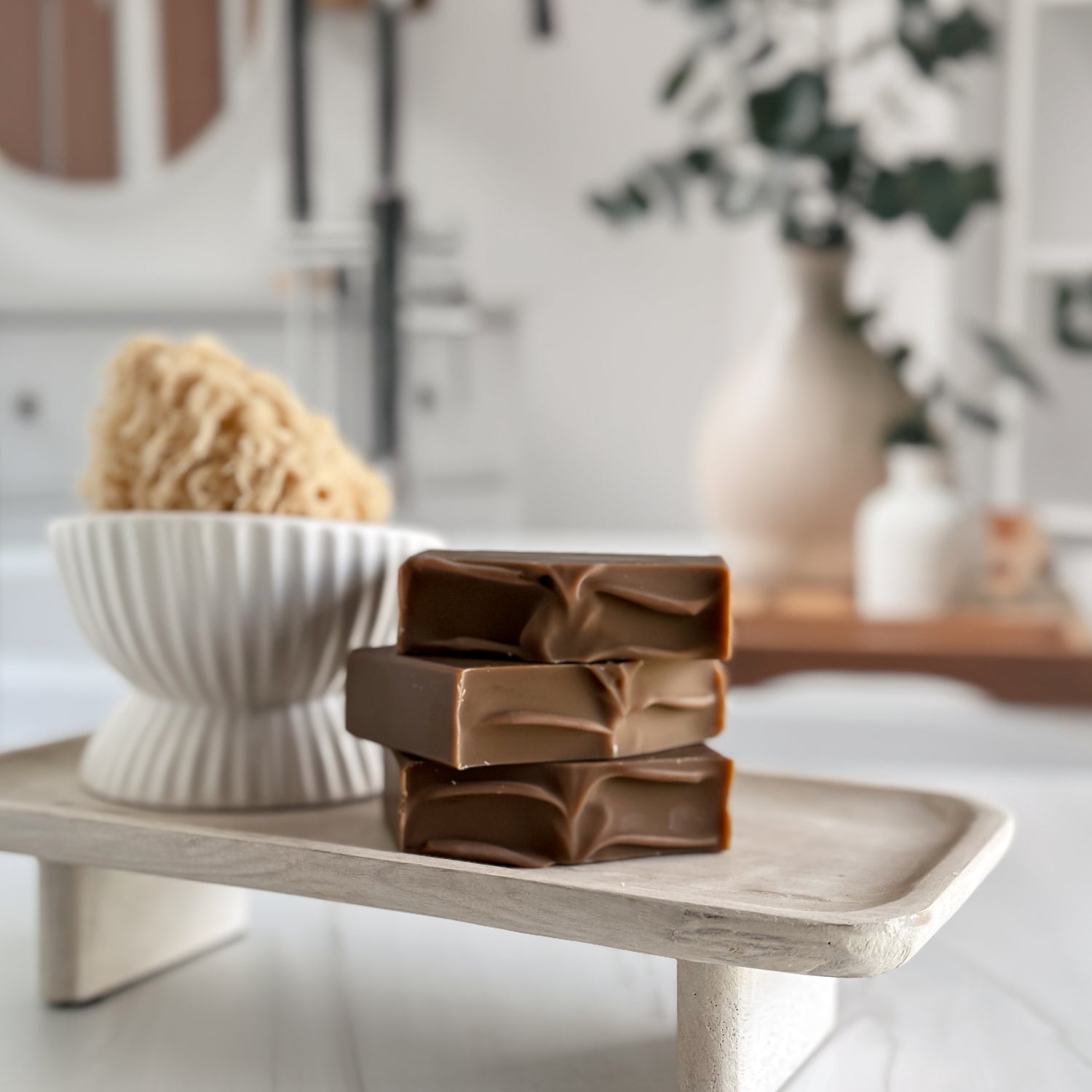 Three bars of soap stacked on a wooden tray with a blurred bathroom background