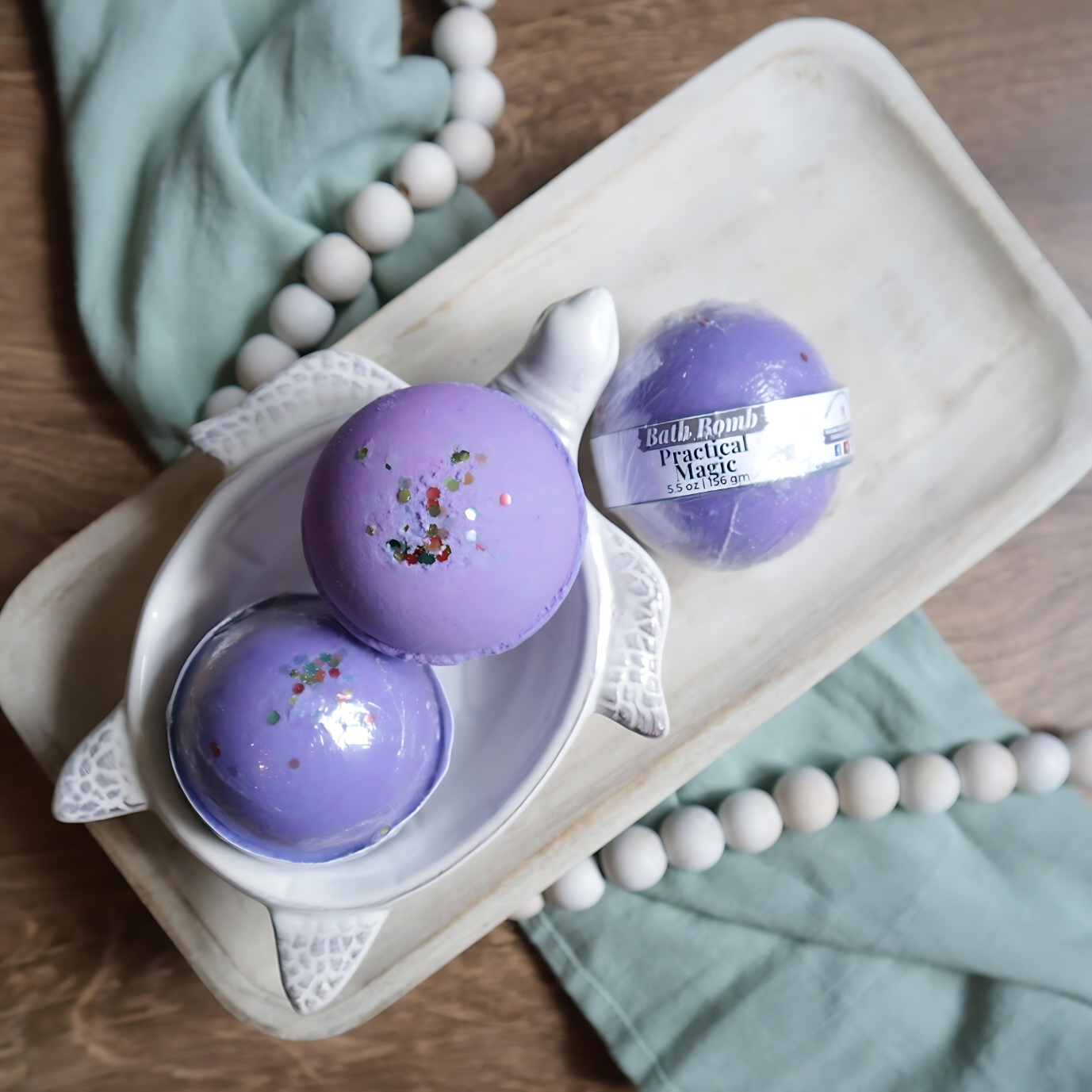 3 practical magic bath bombs are shown sitting on a tray. two are in a soap dish in the shape of a turtle. the bath bombs are purple  in color along with a multi colored glitter on the tops. there is a warm wood base and a green towel with white beads running under the tray and through the picture