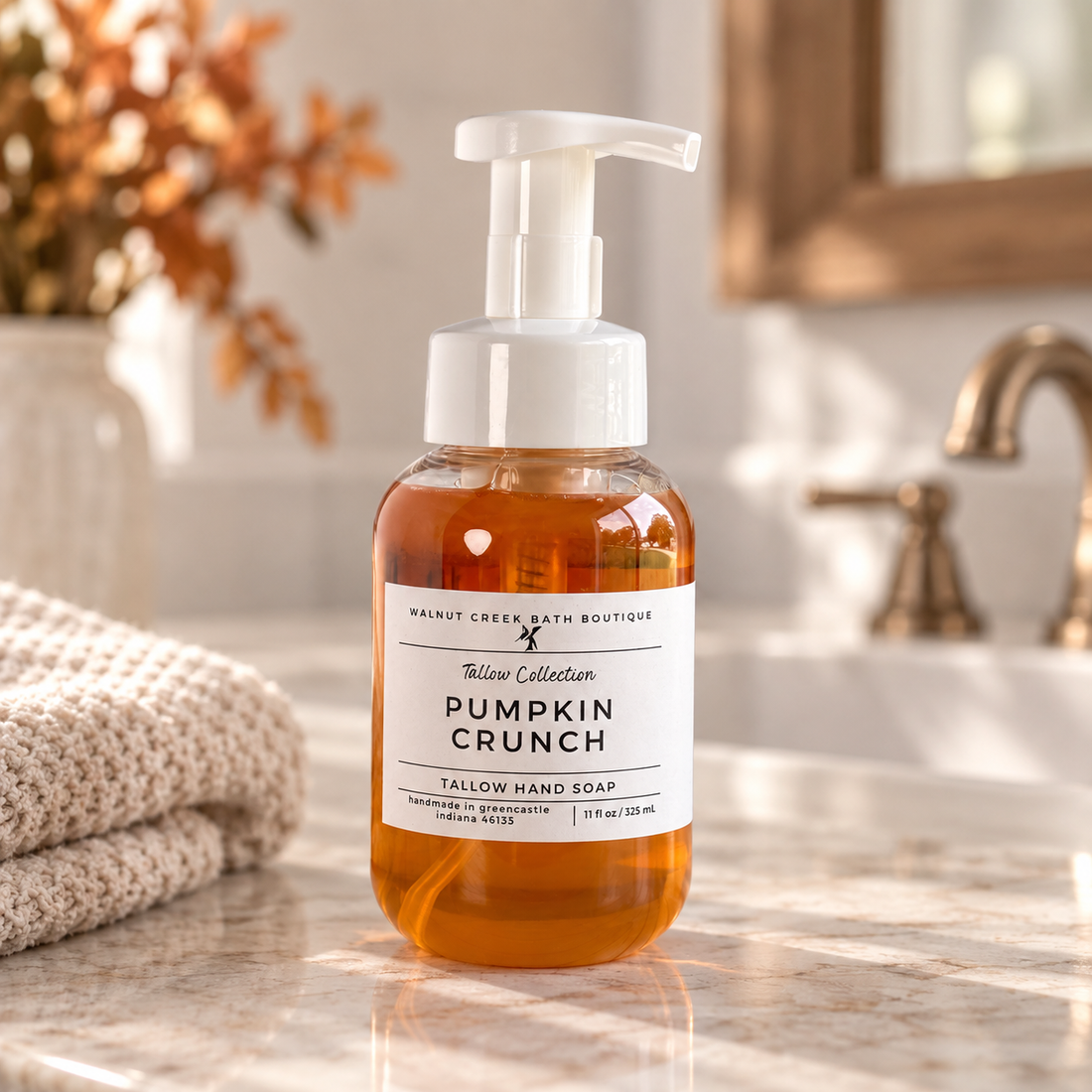 A bottle of Tallow Creek Bath Boutique Pumpkin Crunch Tallow Hand Soap stands on a marble bathroom counter next to a folded towel, with a blurred sink and faucet in the background.