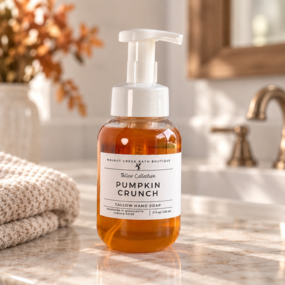 A bottle of Tallow Creek Bath Boutique Pumpkin Crunch Tallow Hand Soap stands on a marble bathroom counter next to a folded towel, with a blurred sink and faucet in the background.