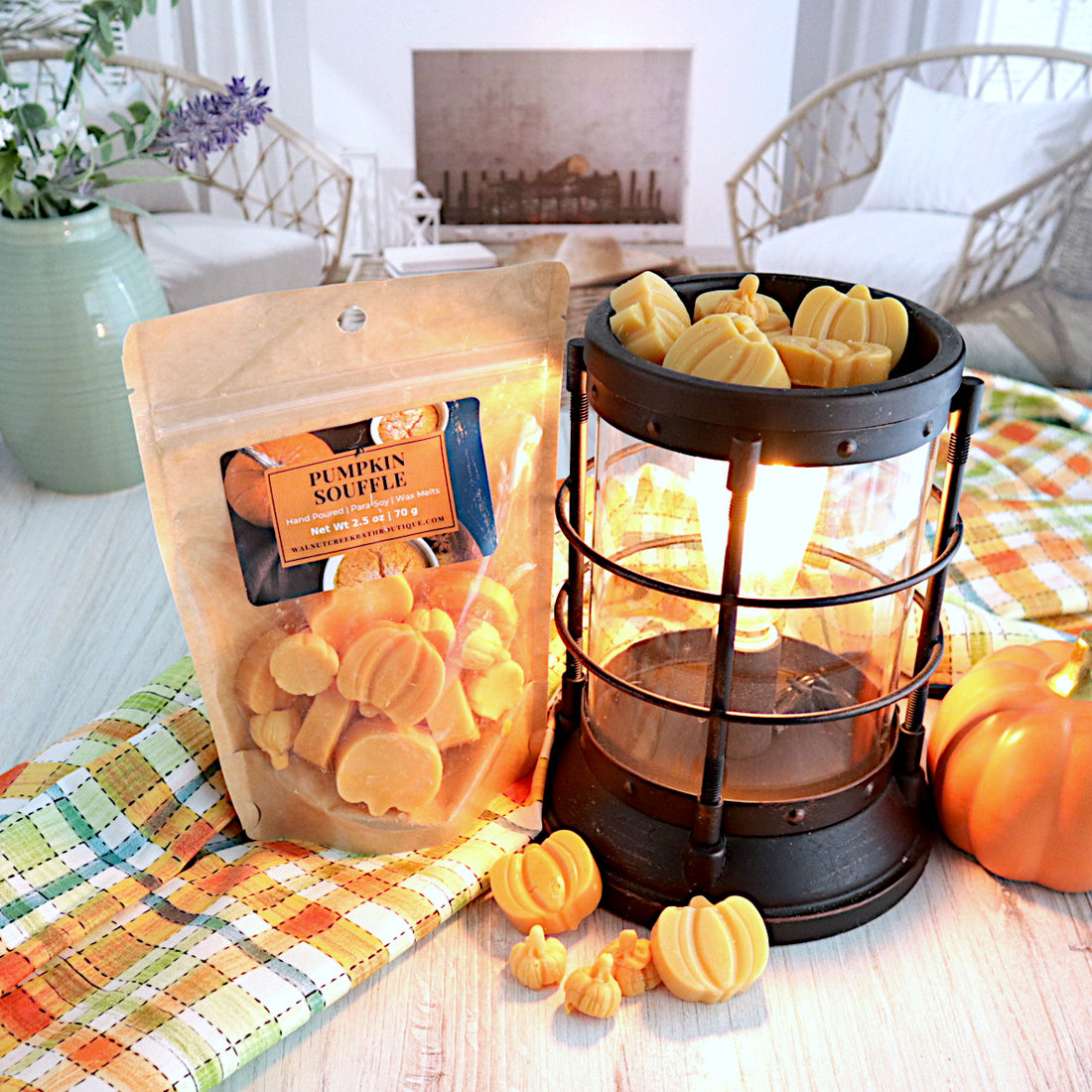 pumpkin souffle wax melt bag standing next to a lit  wax melt burner. there are pieces of wax melts in the burner and also some on the table in front of the burner. the wax is in the shape of large and small pumpkins and is orange om color. there is a plaid fabric in the colors of orange, yellow, green and white that is running through the scene. this is all sitting on a washed out wooden base with a couple of chairs in the background next to a fireplace
