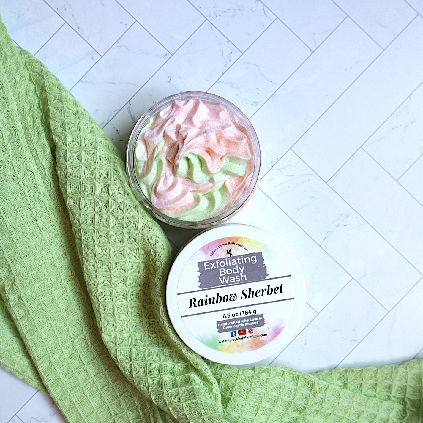 rainbow sherbet scrub is shown with the top off and to the bottom of the jar. inside you will find swirls of pink, orange and green scrub that looks as luscious as it feels. running along side of this is a green fabric with texture.