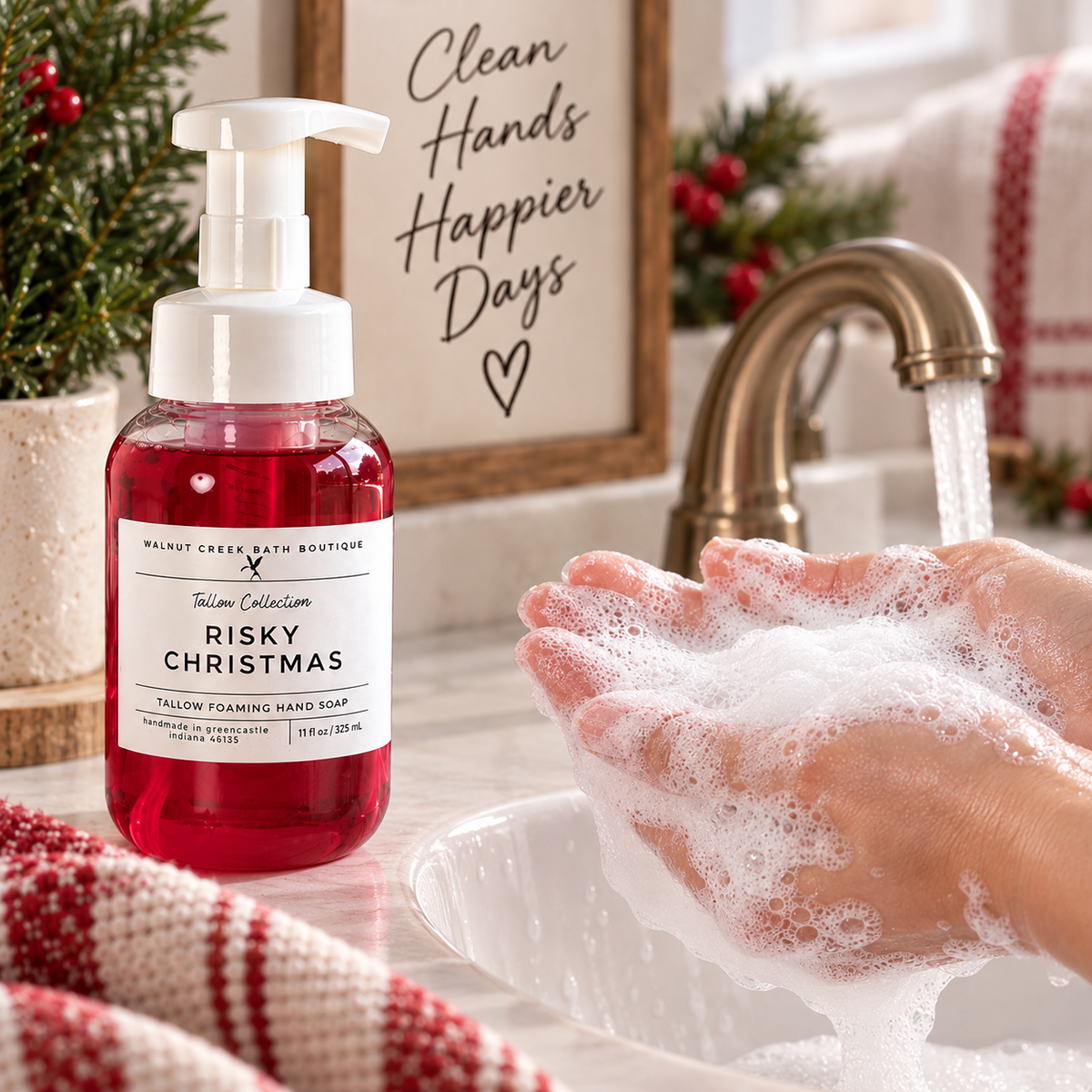 Walnut Creek Hand Boutique Risky Christmas Tallow Foaming Hand Soap bottle next to a sink with soapy hands under a running faucet, with holiday decorations and a sign in the background.