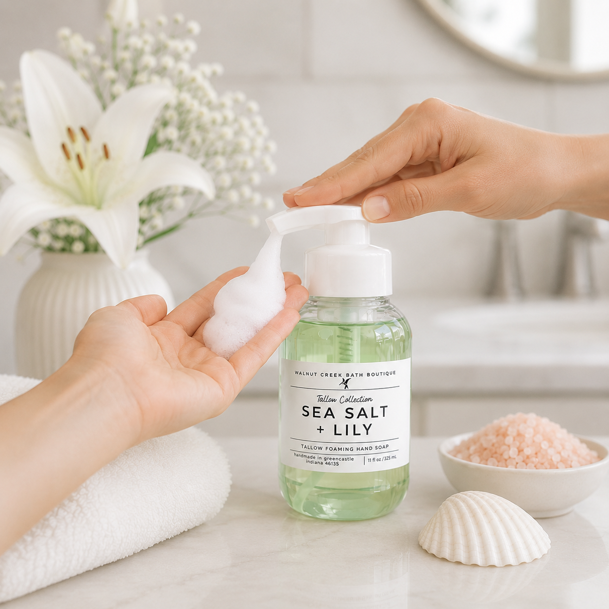 A bottle of sea salt + lily hand wash with a pump dispenser, shown with hands dispensing white foam in a bright bathroom setting with a lily flower, towel, and seashells.