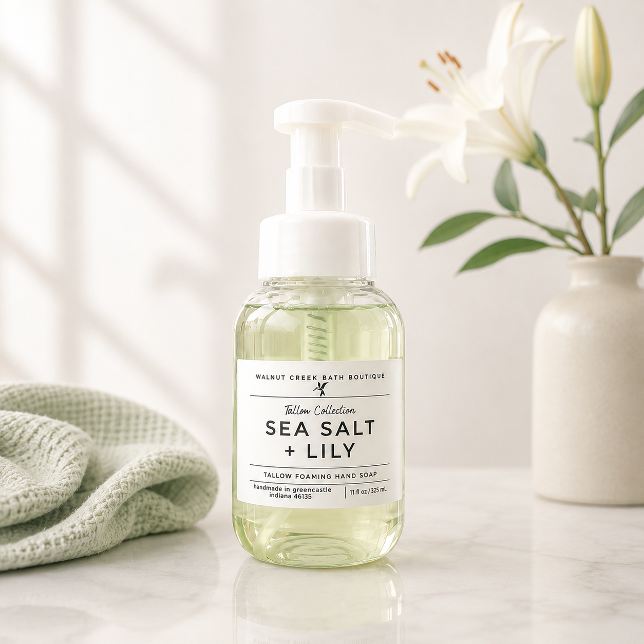 Walnut Creek Bath Boutique Sea Salt + Lily Tallow Foam Wash hand soap bottle on a marble countertop with a green cloth and white lilies in the background.