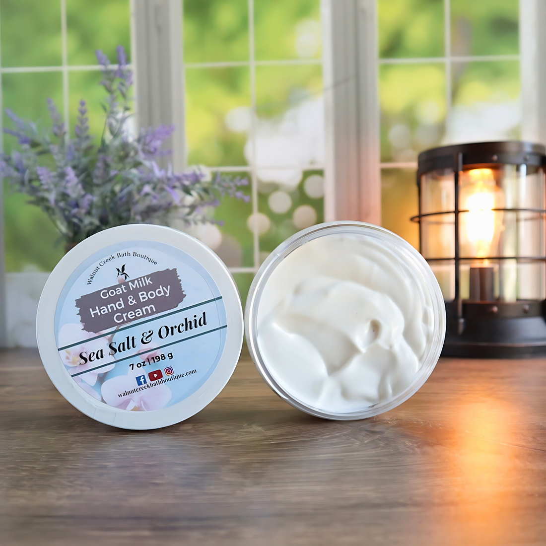 2 sea salt and orchid creams are shown standing on their sides. One to show the label and the other to show the creamy swirly contents. in the back left is a vase with lavender buds and back right is a lit wax burner to add some light. behind all of this is a window with some greenery showing through.