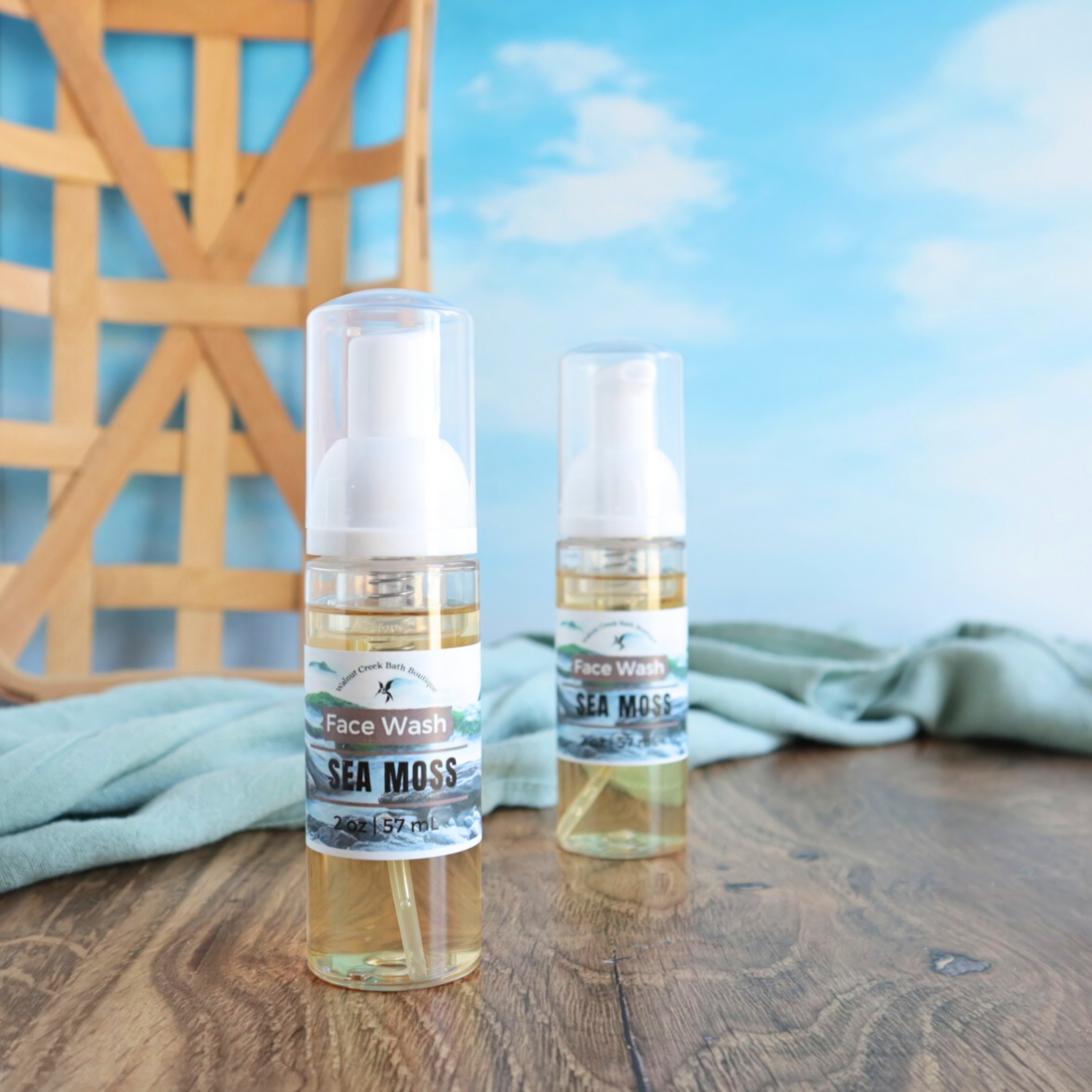 2 sea moss face washes are shown with one more forward than the other. there is a warm brown board base and a green towell running behind the face wash bottles. there is a woven basket in the background with a blue sky with white clouds as a backdrop