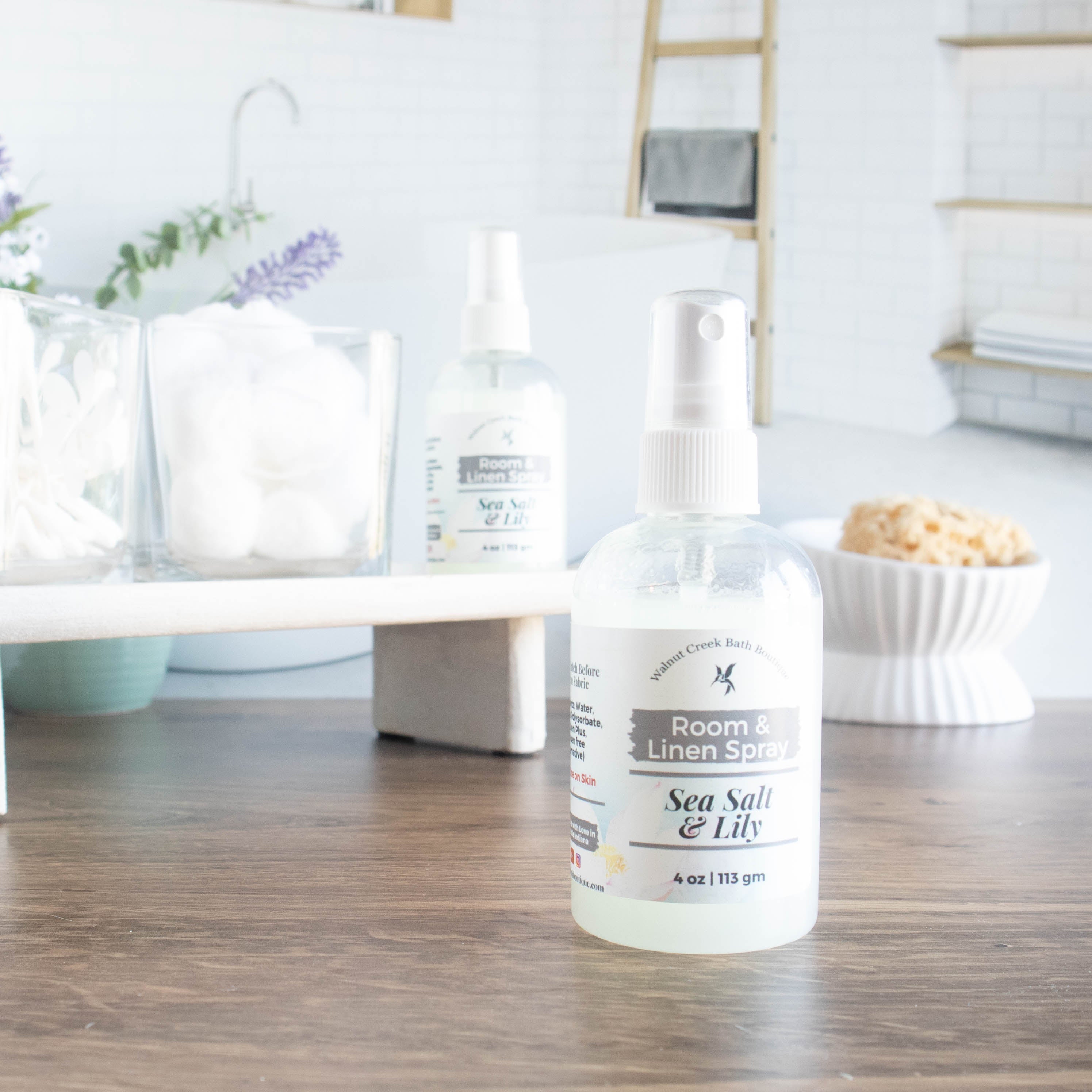 Sea Salt and Lilly room spray is front and center. it is in a squat round bottle with black sprayer. in the background is a tray with some cotton balls and q-tips in jars along with another room spray. in the background is an image of a spa bathroom and a dish with a loofah