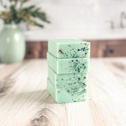 Four stacked rectangular bars of pale mint green soap with dark and red botanical inclusions, displayed on a light wood tabletop with a blurred green vase in the background.