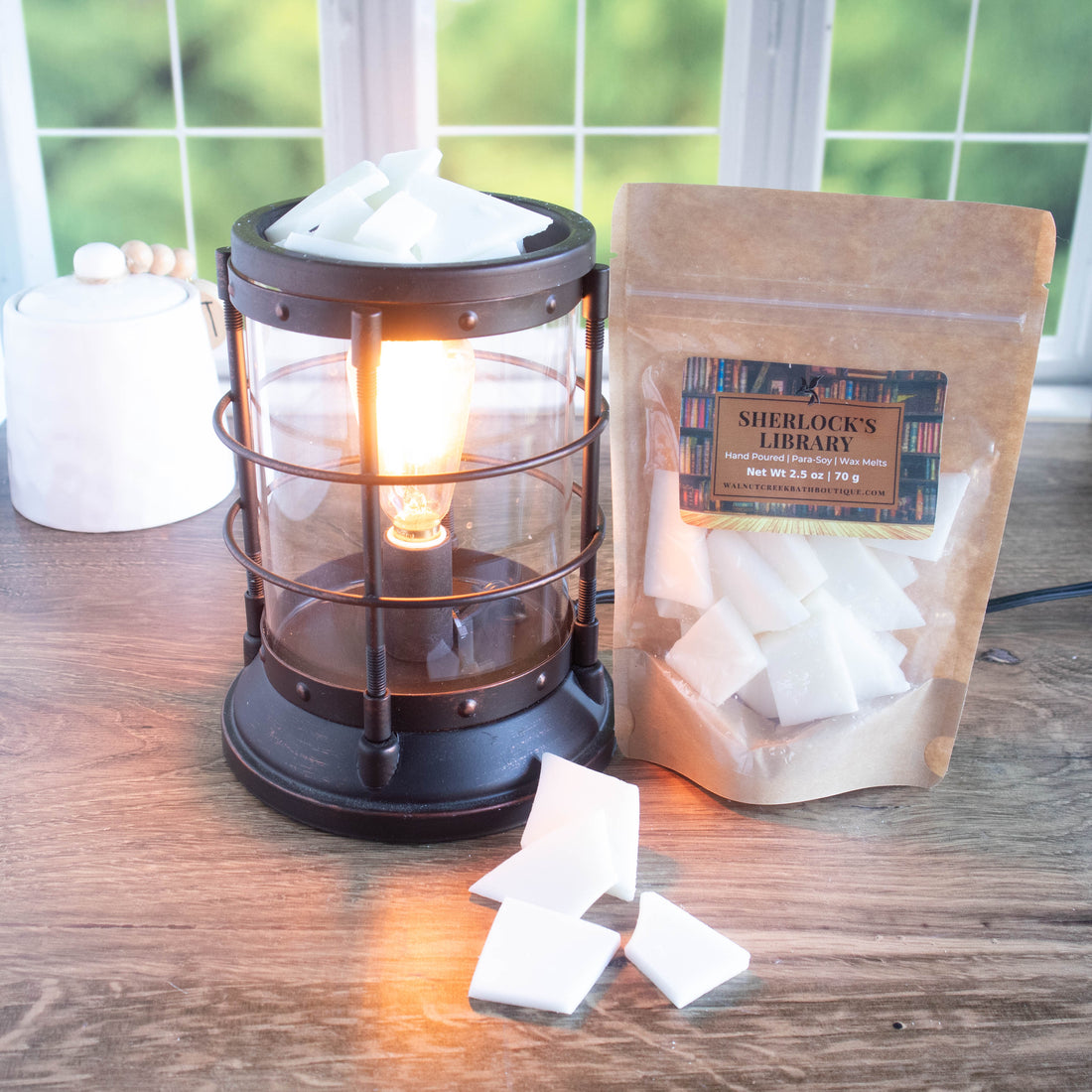 sherlocks study wax melt bag is standing to the right of a wax burner that is bronze in color and turned on. there are white wax pieces in the burner along with a few at the base of it. there is a window in the background showing green leaves.