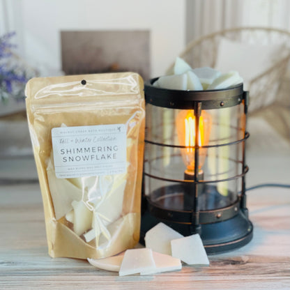 shimmering snowflake wax melt bag is full and standing next to a rustic farmhouse type burner that is on and filled with the white brittle pieces of wax. there are also a few shards of the brittle on the table in front of both to show the size and color of the pieces