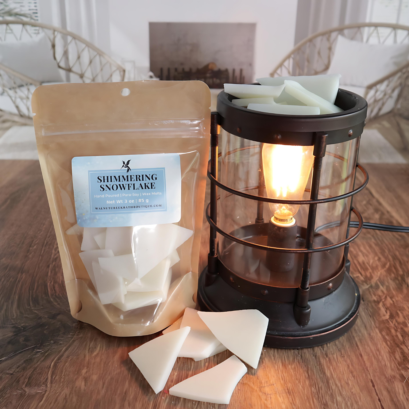 A shimmering snowflake wax melt bag is standing next to a lit bronze wax melt burner. this has the white shards of wax in it and there are some more of these scattered along the base. in the background is a fireplace flanked by 2 wicker chairs.