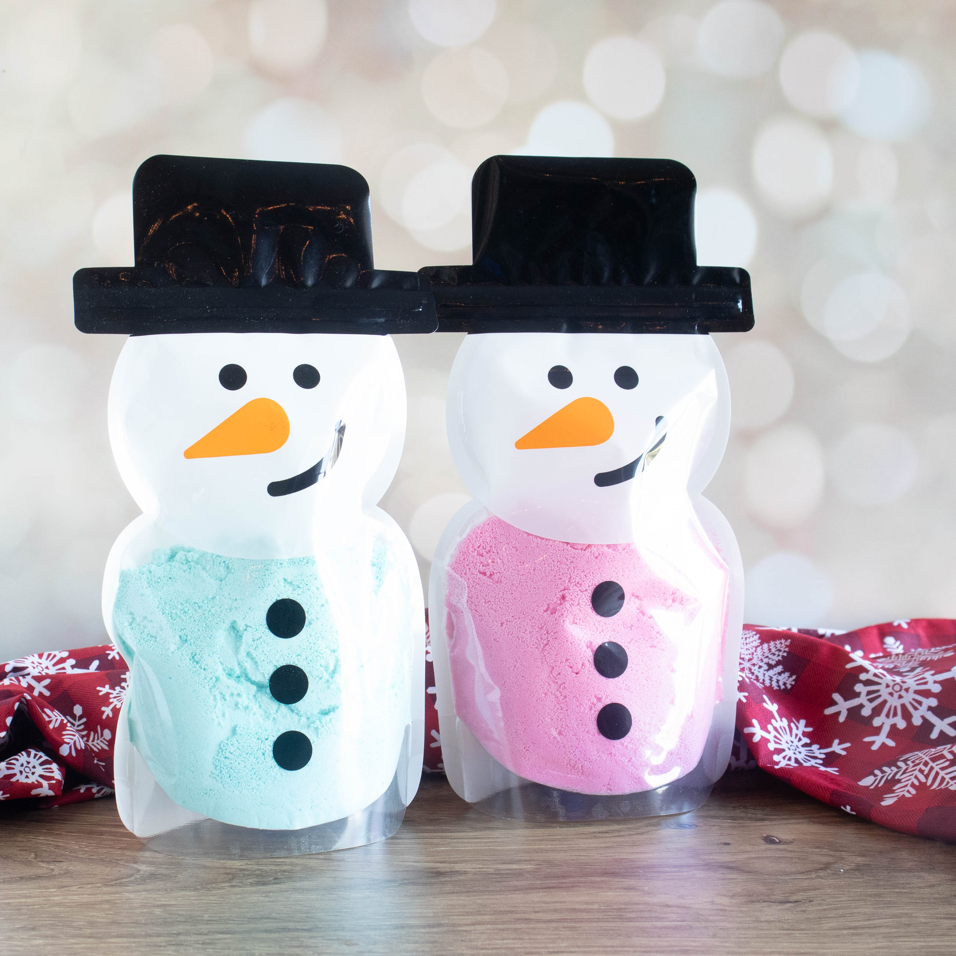 there are 2 bags in the shape of snowmen standing side by side. the bags have a snowman face, hat and buttons to make them super cute! there is green bath bomb powder in the left one and pink in the right one. there is a red fabric running behind the snowmen and it has white snowflakes on it.