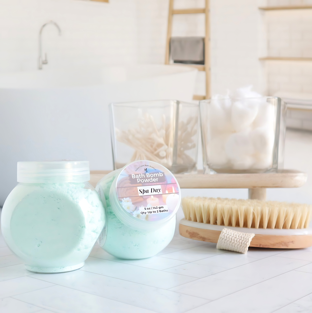 two 5 oz jars of green spa day bath bomb powder in the foreground. One showing the label and another showing the side of the jar with the powder.In the background is a tray with cotton balls and q-tips and a body brush. there is a spa like bath room in the background