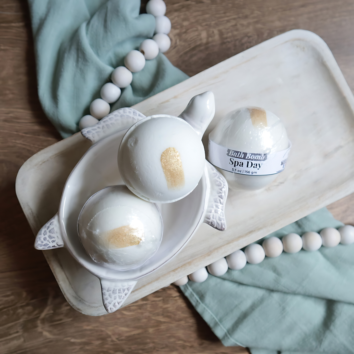 3 spa day bath bombs are shown sitting on a tray. two are in a soap dish in the shape of a turtle. the bath bombs are white in color with a gold swash to give a pretty interest. there is a warm wood base and a green towel with white beads running under the tray and through the image