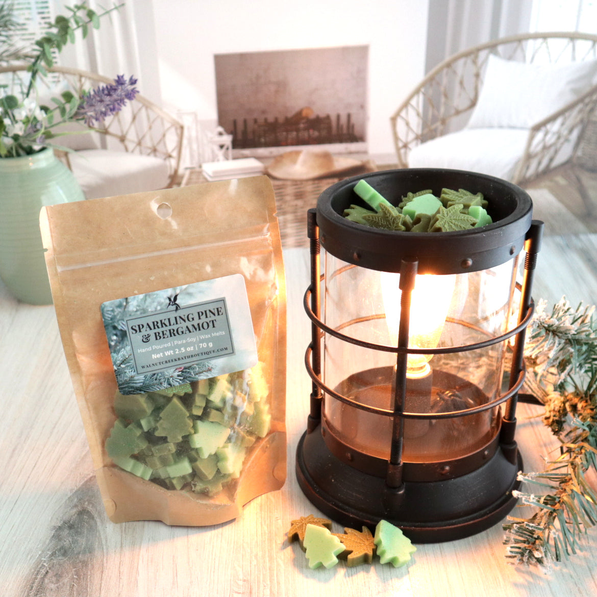 A sparkling pine and bergamot wax melt bag is standing next to a lit bronze wax melt burner. this has the several different green colored bits of wax in the shape of trees and leaves in it and there are some more of these scattered along the base. in the background is a fireplace flanked by 2 wicker chairs.