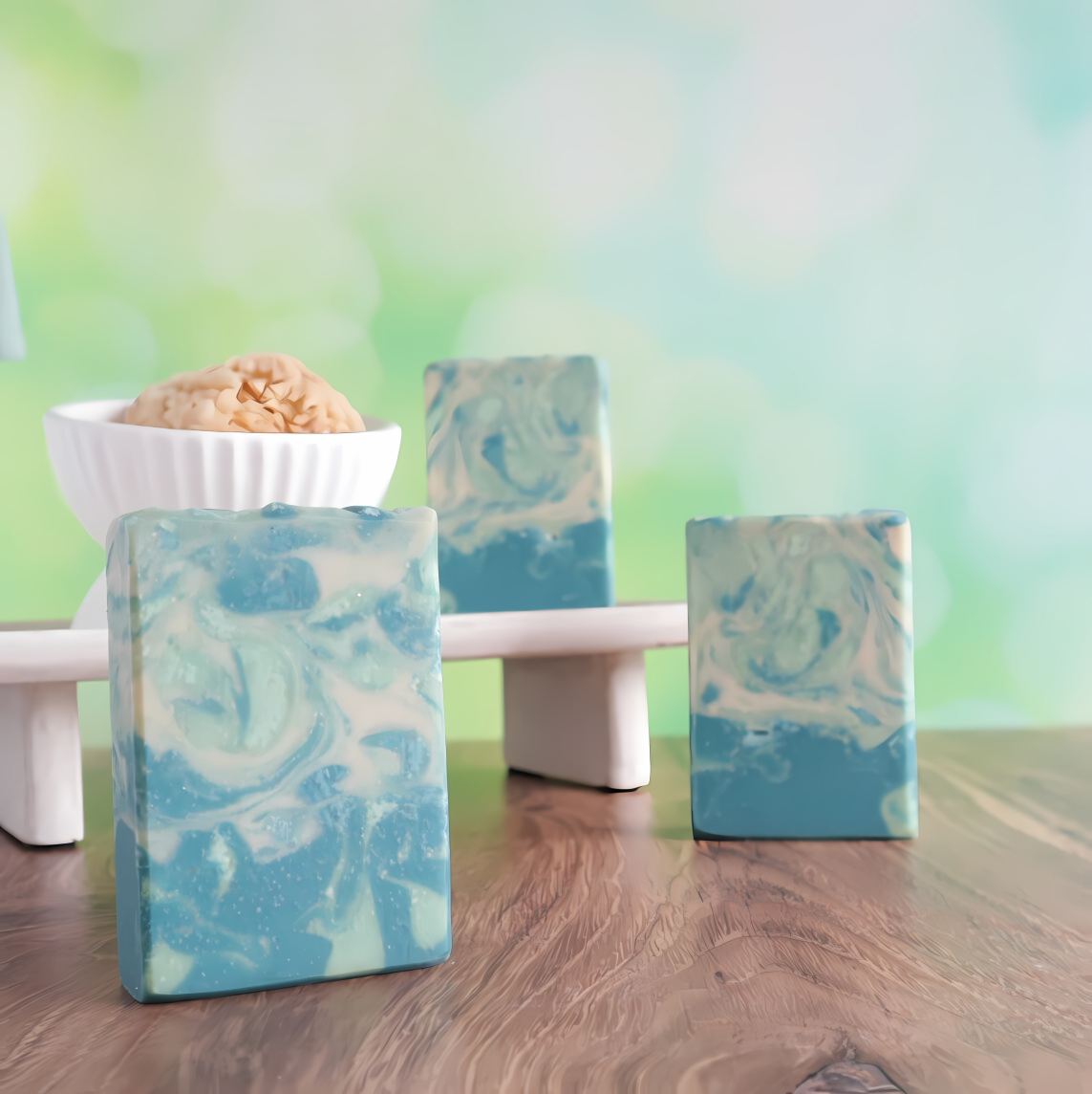 3 sparkling pine soaps are shown. they have blue, green and cream swirls all throughout them. one soap is standing front left. there is one a little back to the right and the 3rd is on a tray behind these two. on the tray is a white soap dish with a loofah peeking out. the background is a green ombre to creamy white