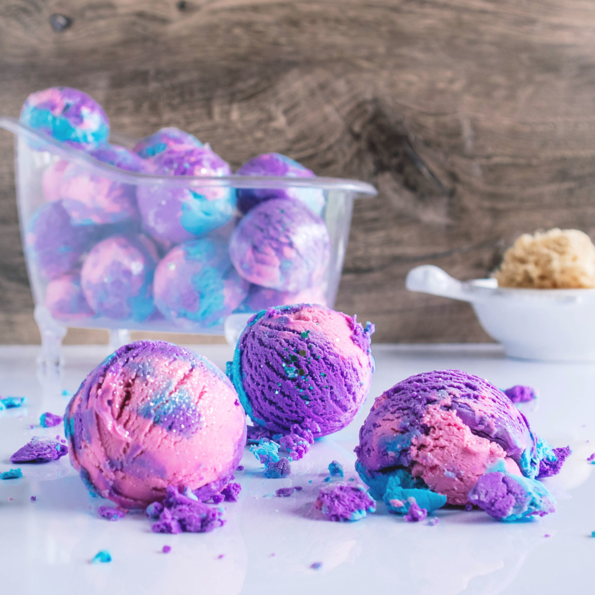 3 solid bubble bath, pink blue and purple with glitter on top are sitting in front of a clear bath tub full of more solid bubble baths.  there is a sprinkling of broken up bubble bath crumbles laying all around