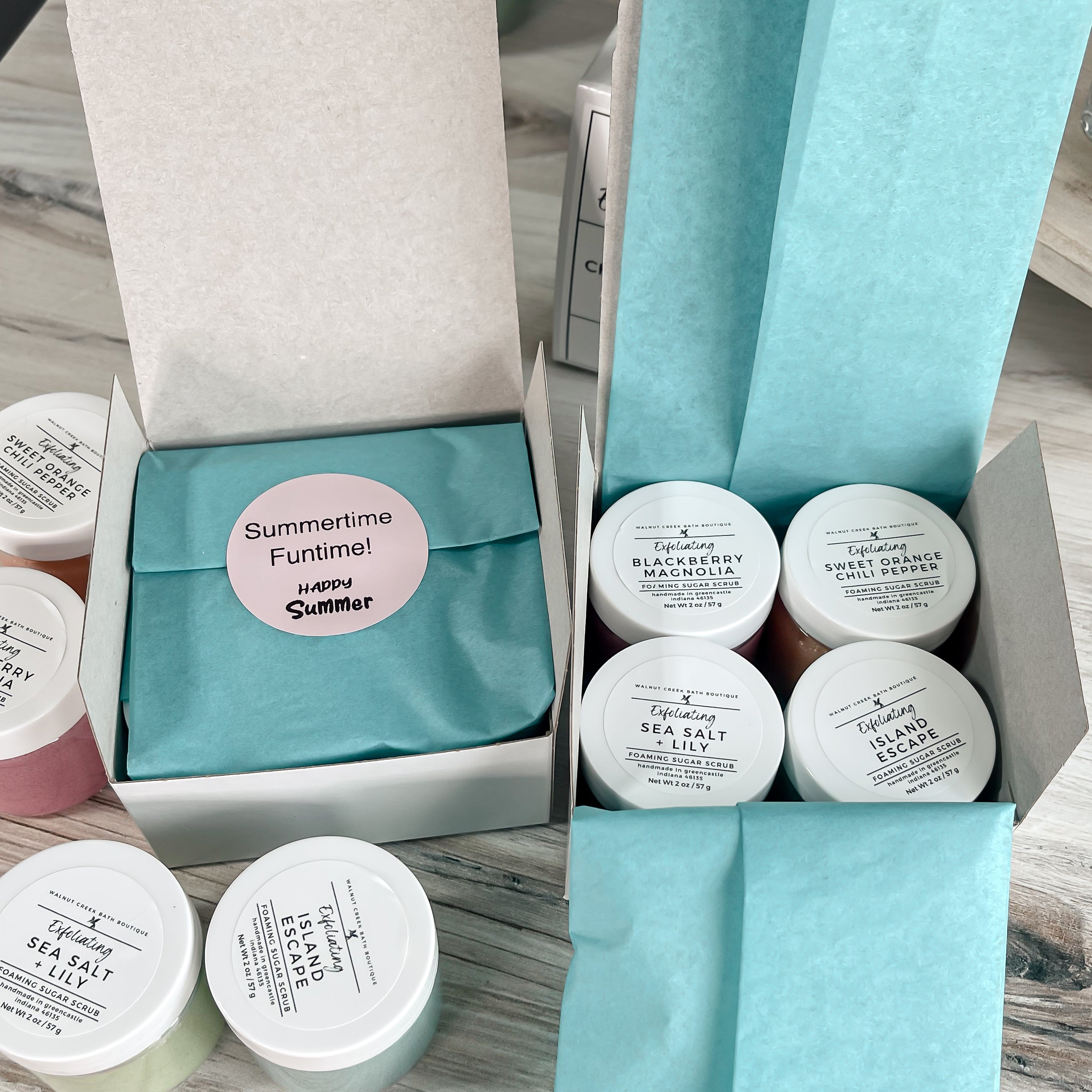 Set of small containers with lids, with one box open to show sticker and one option to show tissue paper open, on a wooden surface.