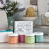 Four small jars of bath scrubs with a sign indicating the brand and collection name, placed on a wooden surface.