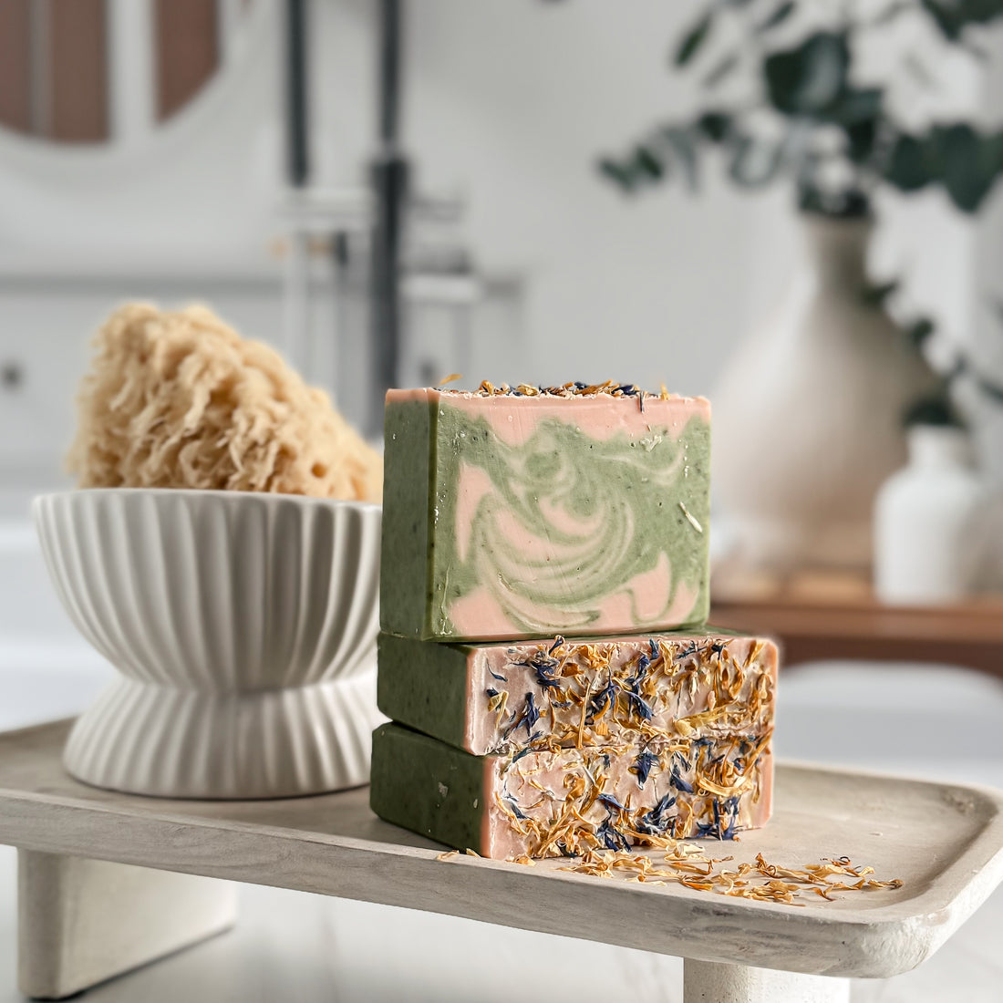 Three soap bars with natural ingredients on a wooden tray, with a bowl and plant in the background.