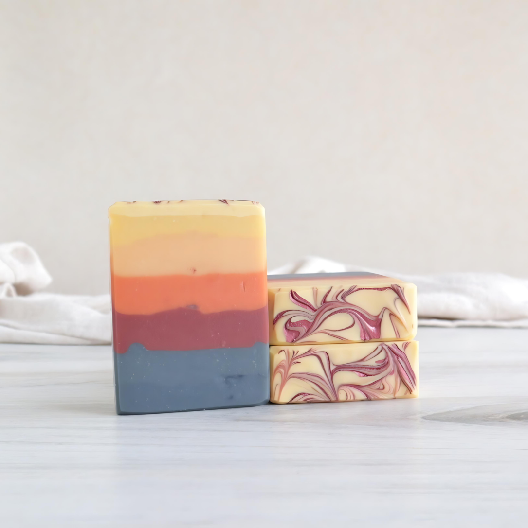 sunset coast soaps are shown, one is standing tall showing the layers that start dark blue at the bottom, then go up to rust red, then orange and finally a yellow. there are 2 bars next to this laying flat showing the tops. the tops have yellow soap with a very pretty red swirl throughout. the background is tan and there is a tan colored towel in the back as well.