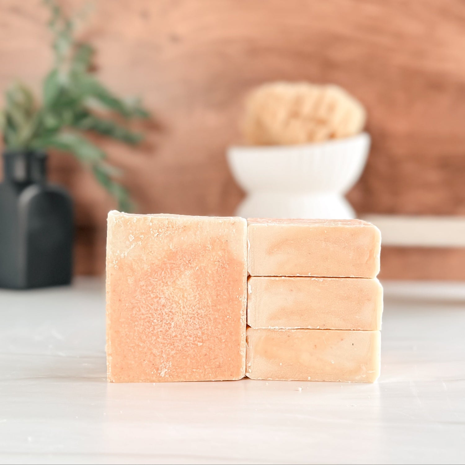 Three bars of soap stacked on a white surface with a wooden background and plant.