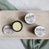 there are 3 tallow face cream jars on a tray.  one of the jars are open to show the pretty yellow color of the cream. there is a sage green towel running underneath and on a diagonal the tray. the base is a herringbone in marbal.