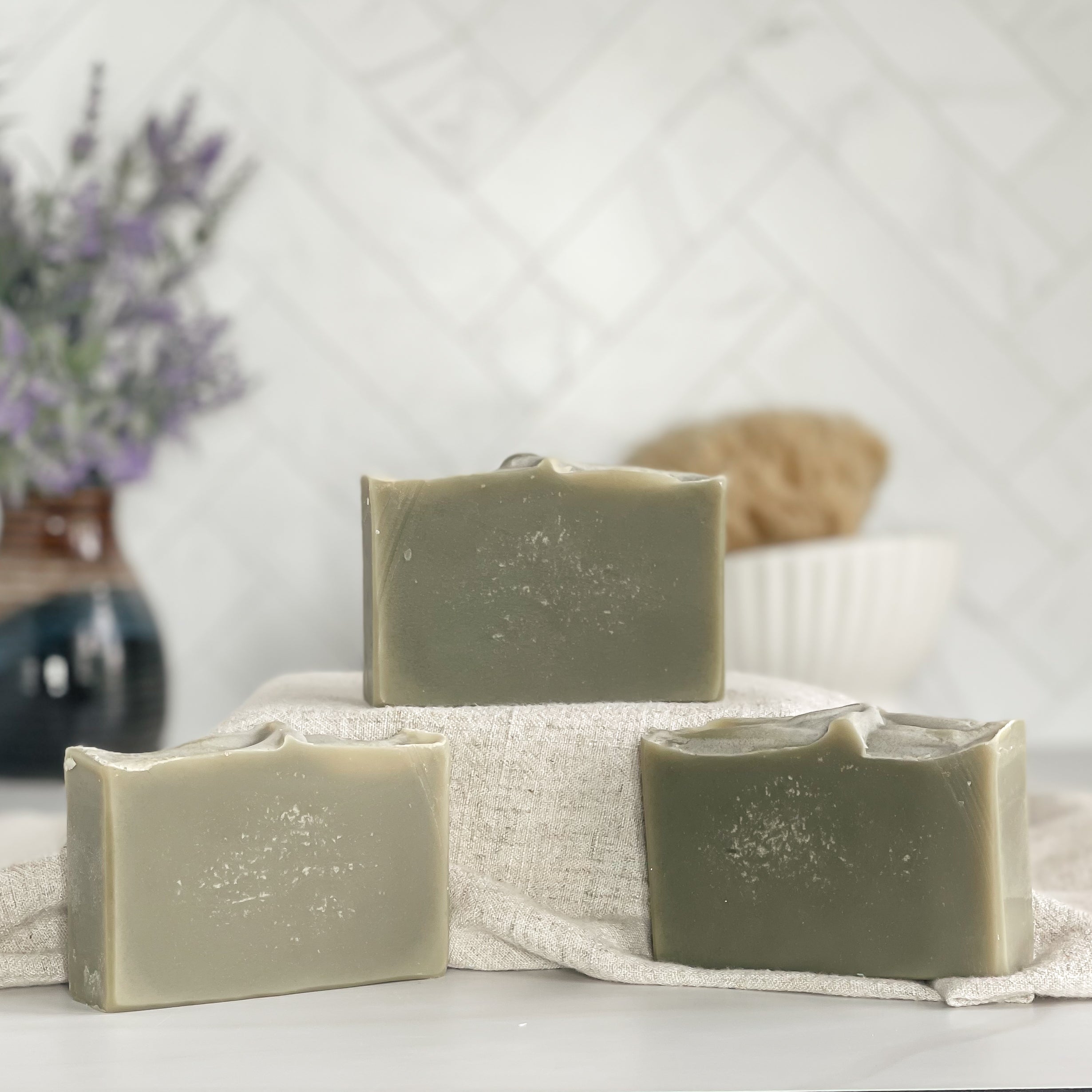 three green oasis soaps are shown with one on a tan covered dias along with a loofah in a dish in the background