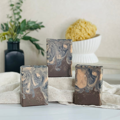three indian sandalwood soaps are shown with one on a tan covered dias along with a loofah in a dish in the background