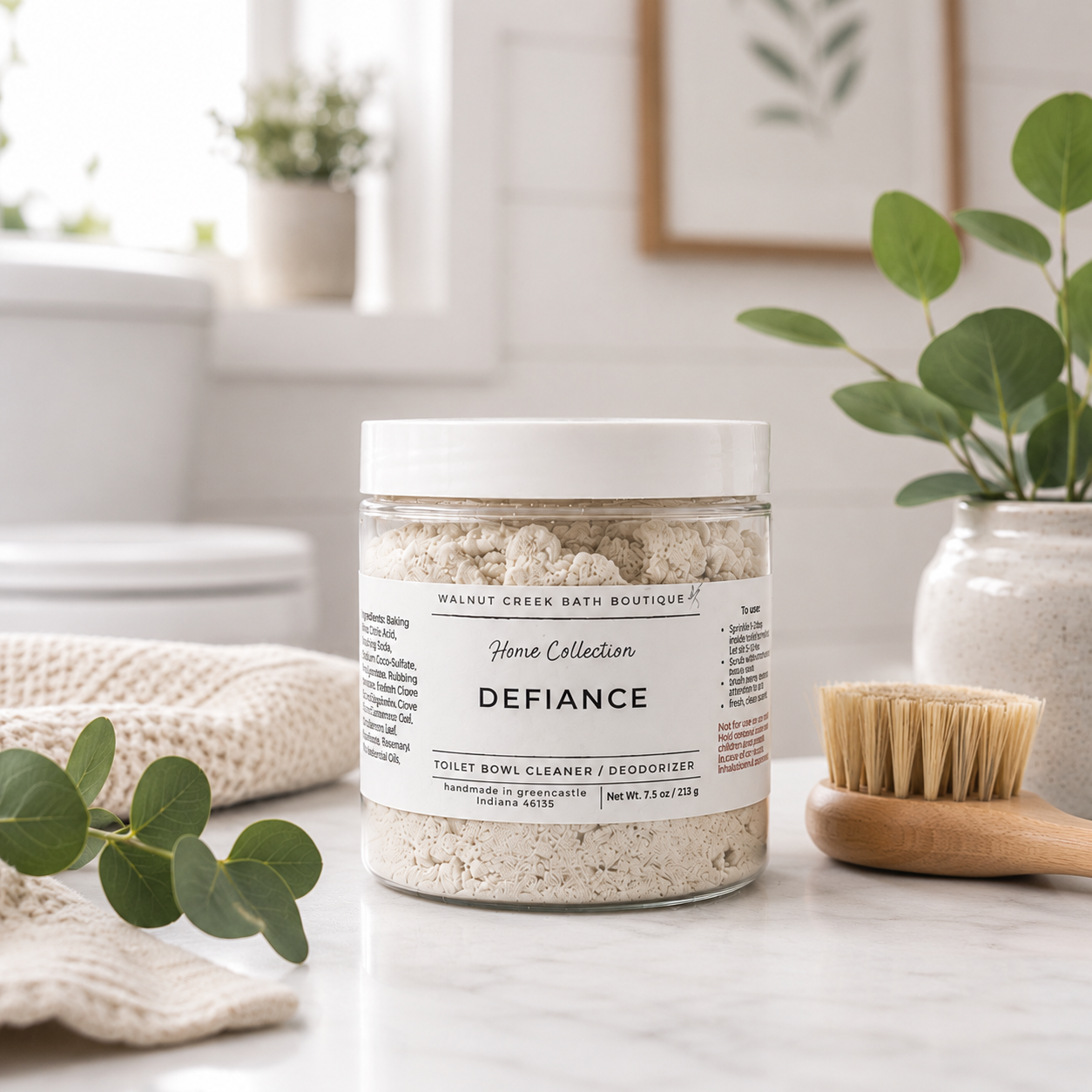 A clear plastic jar of walnut creek bath boutique Defiance toilet bowl cleaner and deodorizer sits on a marble countertop with a wooden brush and eucalyptus leaves in a bright bathroom setting.