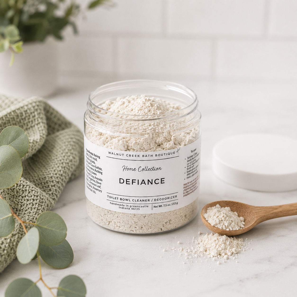 A clear plastic jar of Home Collection Defiance Toilet Bowl Cleaner by Walnut Creek Bath Boutique, filled with a light beige powder, styled on a white marble surface with a wooden spoon, a white lid, and green eucalyptus leaves.