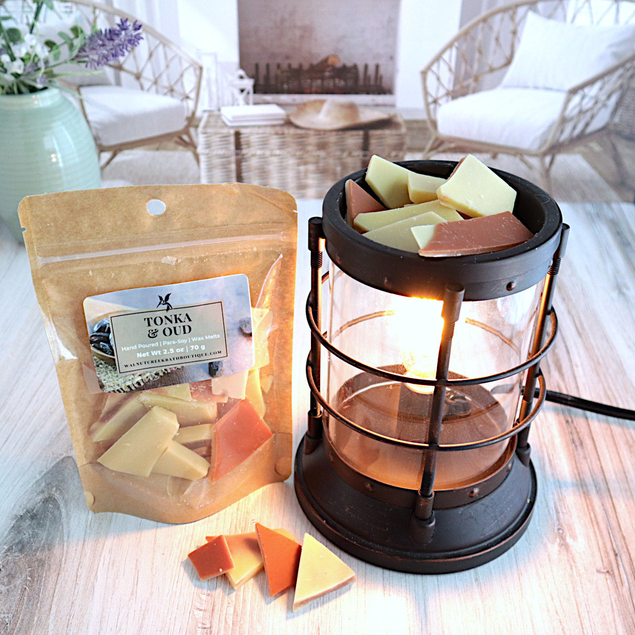 tonka and oud wax melt bag is standing next to a lit burner full of wax pieces. the wax is in small shard like pieces, tan in color with some brown on top. there a some pieces on the table in front of the bag and burner as well. this is all sitting on a washed out wooden base with a couple of chairs in the background next to a fireplace