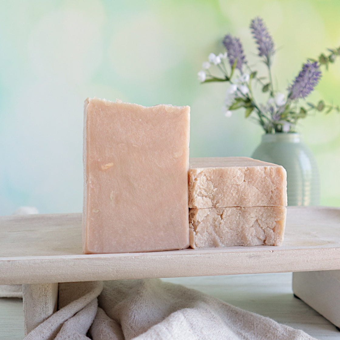 there are 3 rainbow sherbert soaps on a tray. they have a tan color and rustic appearance. 1 is standing tall while 2 more are next to it laying flat to show the tops. there is a soft green backdrop and a vase in the back right with some flower buds.