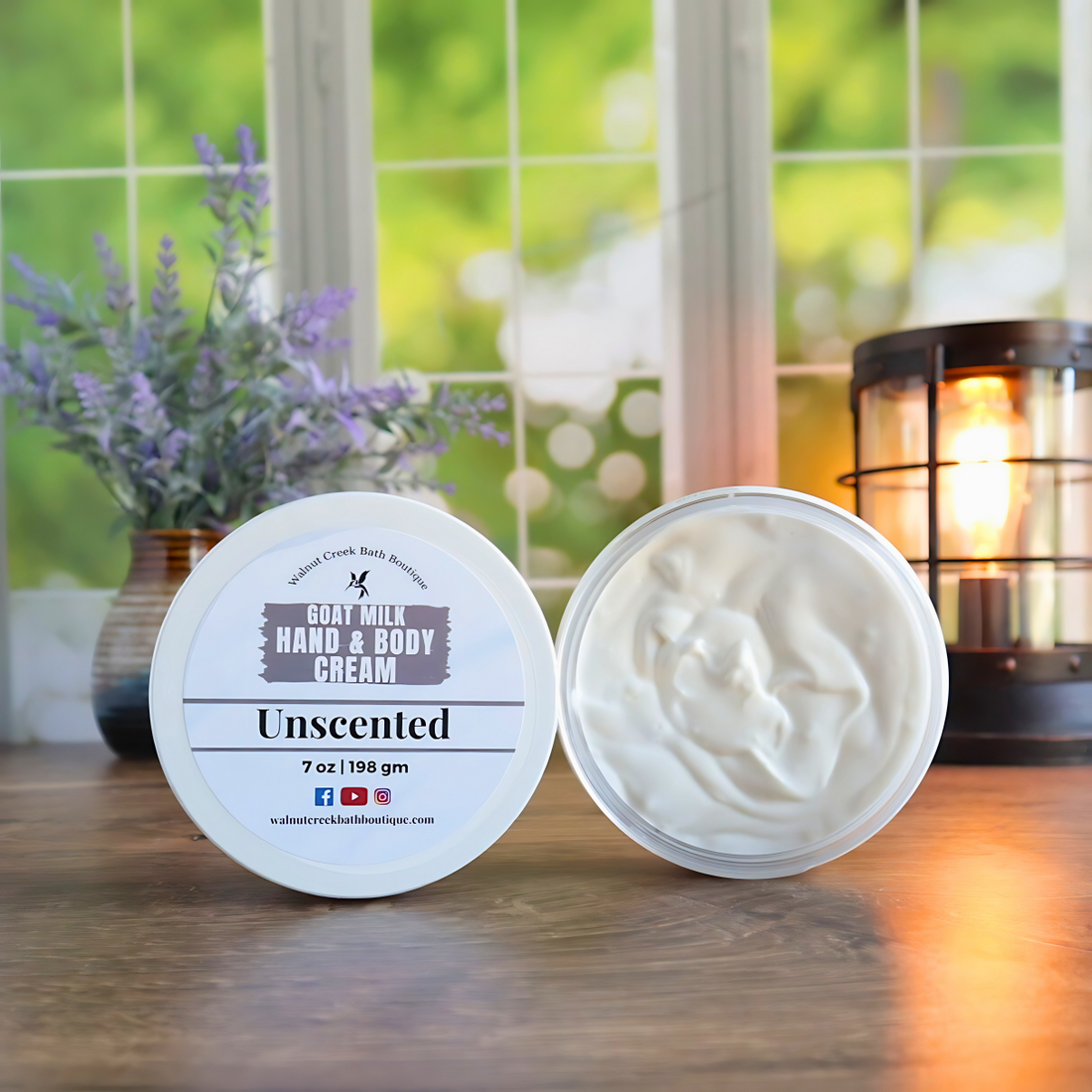 2 unscented creams are shown standing on their sides. One to show the label and the other to show the creamy swirly contents. in the back left is a vase with lavender buds and back right is a lit wax burner to add some light. behind all of this is a window with some greenery showing through.