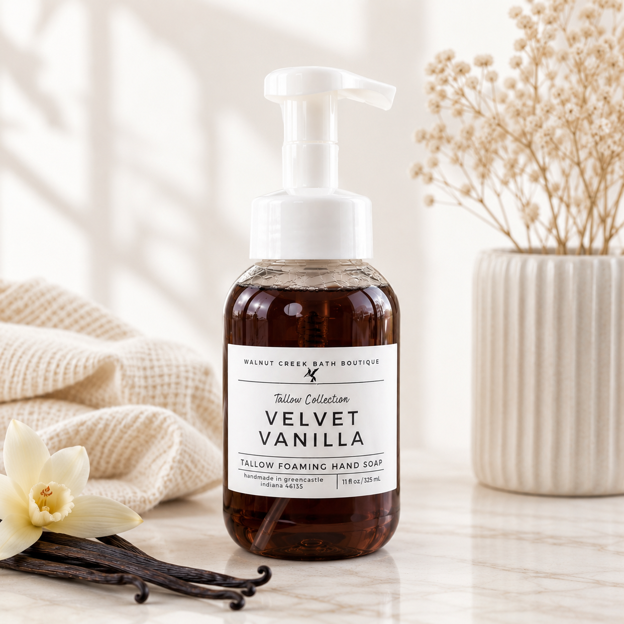 A bottle of Walnut Creek Bath Boutique Velvet Vanilla Fallow Foaming Hand Soap stands on a marble surface with a beige towel, vanilla beans, and dried flowers in the background.