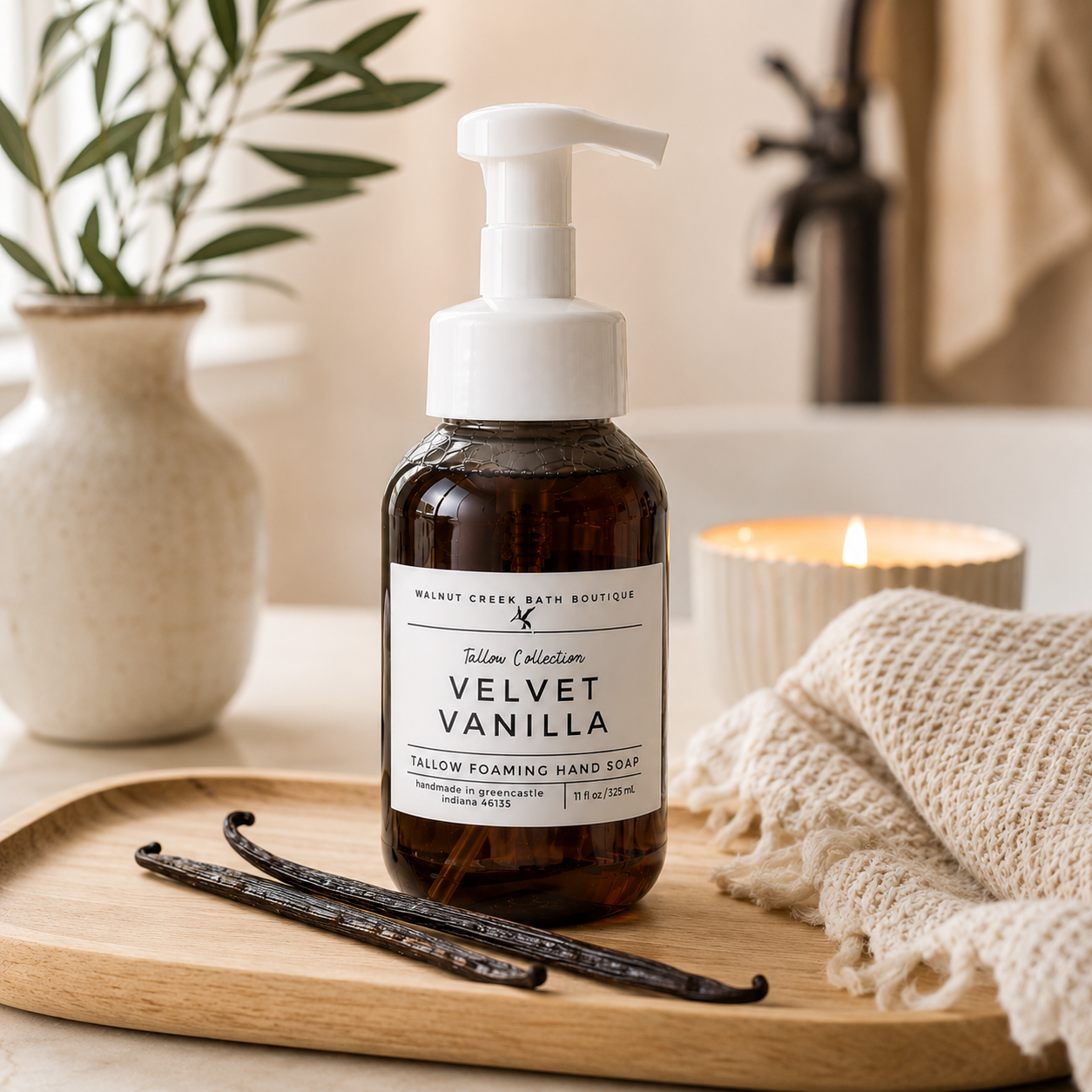 A bottle of Mount Green Bath Boutique Vanilla Collection Velvet Vanilla follow foaming hand soap sits on a wooden tray in a bathroom setting, surrounded by vanilla beans, a knit towel, and a lit candle.