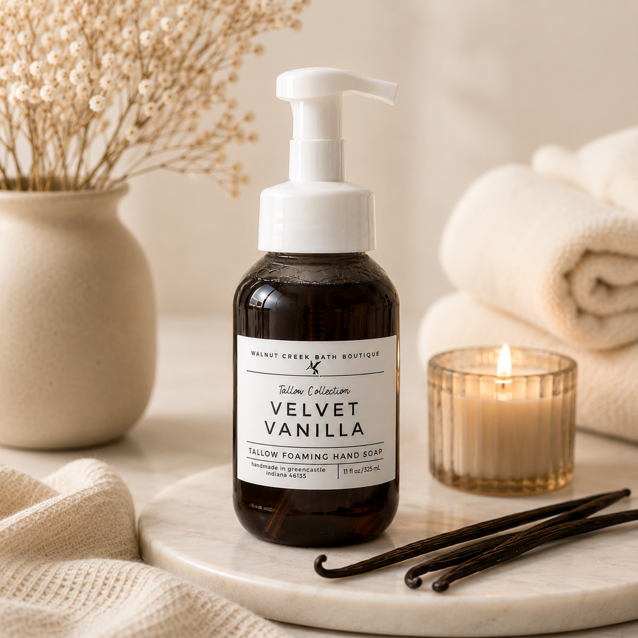 A bottle of Walnut Creek Bath Boutique Fallow Collection Velvet Vanilla foaming hand soap sits on a marble tray, surrounded by a lit candle, vanilla beans, and folded towels in a warm, cozy setting.